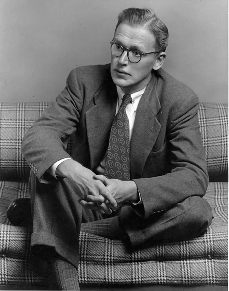 Person with glasses sitting on a plaid sofa, wearing a suit and patterned tie, hands clasped, looking to the side thoughtfully.