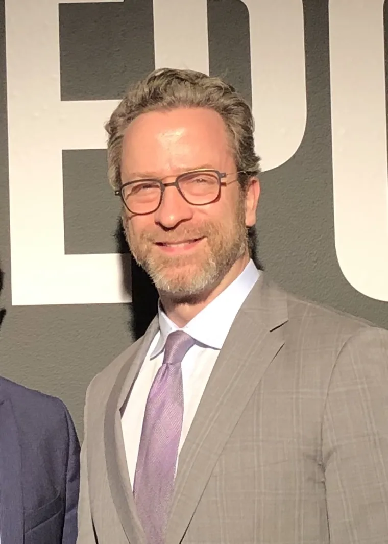 Person with glasses and beard, wearing a light gray suit and lavender tie, stands against a backdrop with partial text visible.