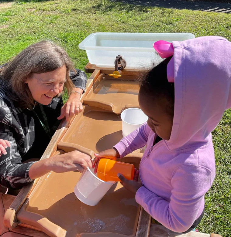 Adult and child playing with water and containers at an outdoor play station. Child wears a pink hoodie; adult smiles and engages in the activity.