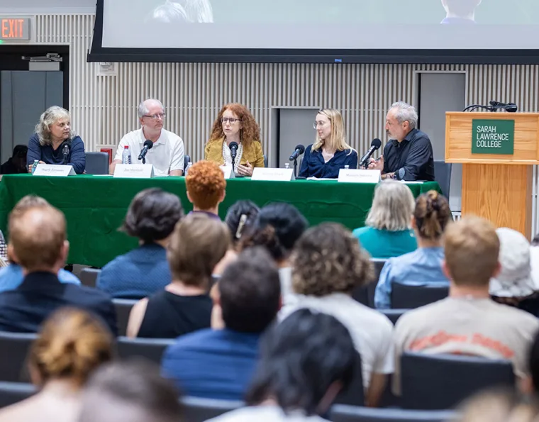 Panel of five people speaking at a table with microphones in front of an audience. Sign on podium reads "Sarah Lawrence College."