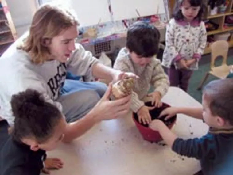 Person showing children how to plant seeds. One child is holding soil, while others watch and join in the activity.