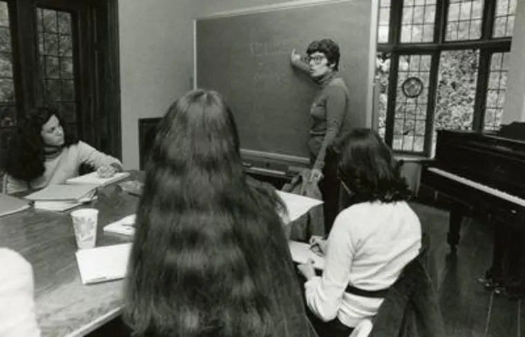 Person writing on a chalkboard in a classroom while others sit around a table taking notes. A piano is in the background near a window.