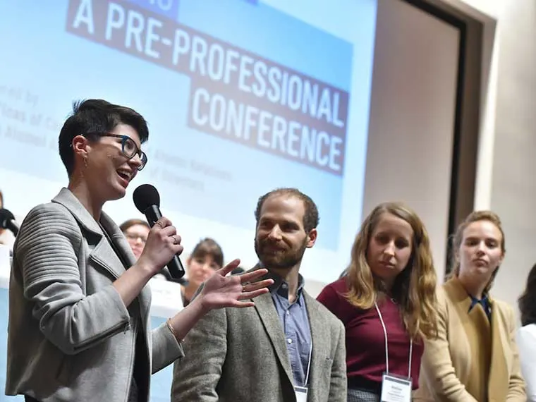 Person with short hair speaks into a microphone at a conference. Three others stand nearby, looking on. A presentation screen is in the background.