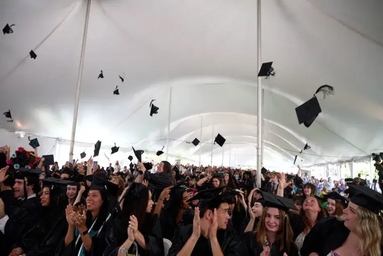 Graduates in caps and gowns celebrate under a large tent, tossing caps in the air, with an audience clapping and cheering.