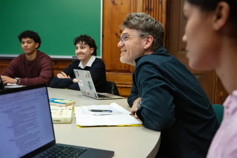 Four people sitting around a table with laptops and books, engaged in a discussion. A green chalkboard is visible in the background.