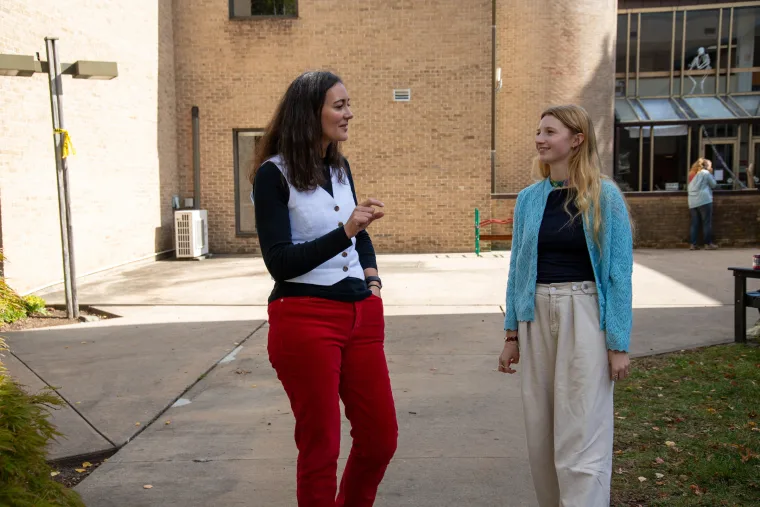 Two people are talking and walking outside near a brick building. One is wearing red pants, and the other is wearing a blue cardigan.
