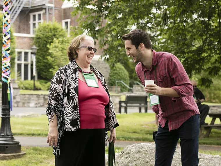 Two people smiling and talking outdoors, one wearing sunglasses and a floral top, the other holding a cup, with trees and a building in the background.