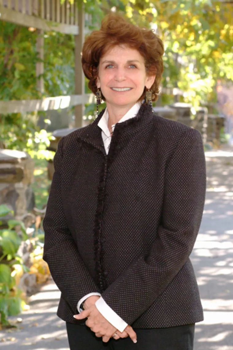 Person in a dark patterned jacket smiling outdoors, with sunlight filtering through green foliage in the background.