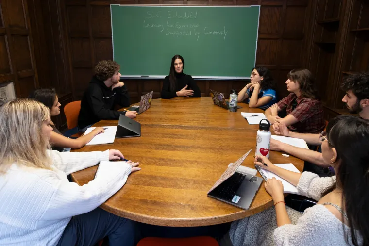 Eight people sitting around a large wooden table in a classroom, engaging in discussion. A chalkboard in the background has writing on it.