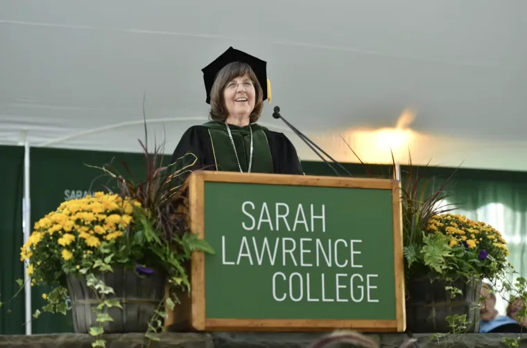 Person in academic regalia speaks at a podium with "Sarah Lawrence College" sign, surrounded by flowers.