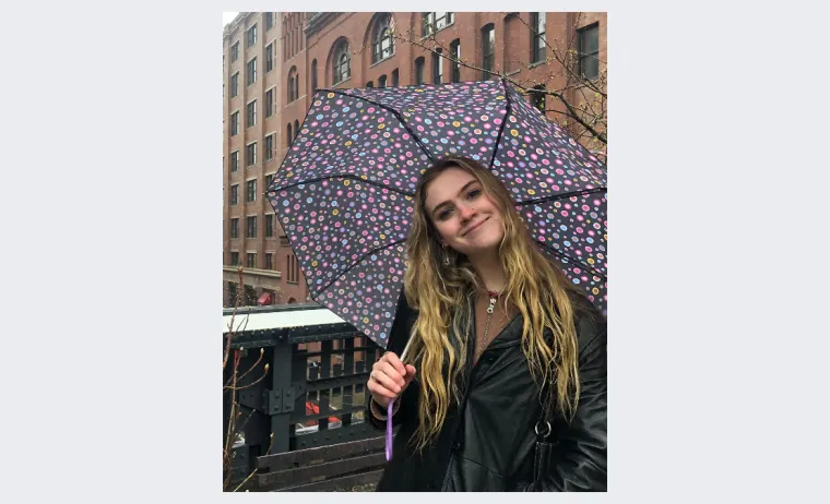 Person with long hair holding a polka dot umbrella, standing on an urban street with brick buildings in the background.