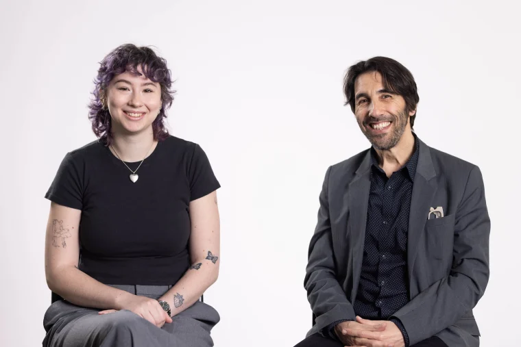 Two people sitting and smiling, one with purple hair and tattoos, the other in a suit with dark hair, against a plain white background.