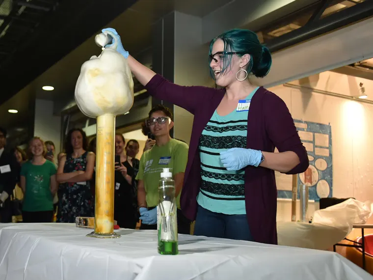 Person with turquoise hair demonstrating a chemical reaction on a table, surrounded by crowd, wearing gloves and a name tag.