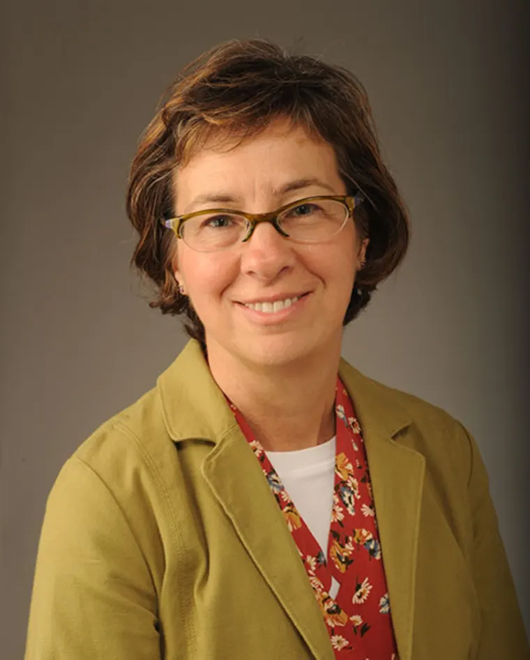 Person with short brown hair and glasses, wearing an olive blazer over a patterned red shirt, smiles at the camera.