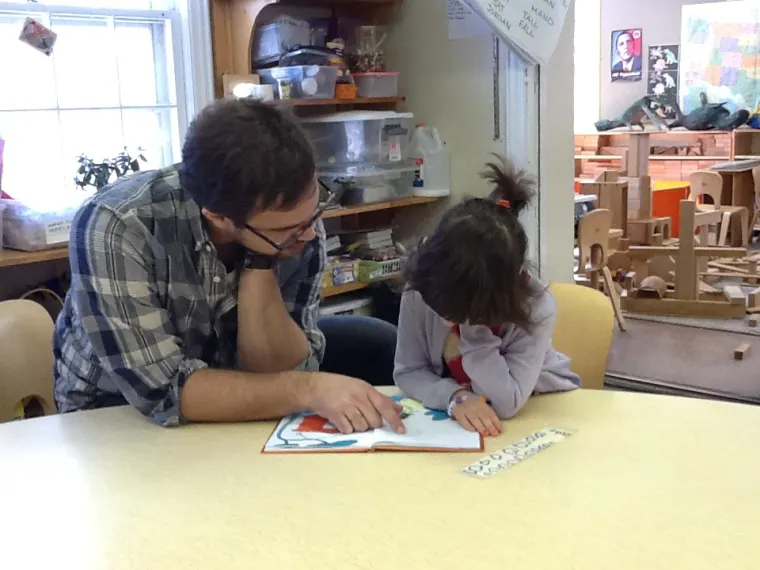 Two people sitting at a table in a classroom, reading a book together. Shelves with supplies and a bright window are in the background.