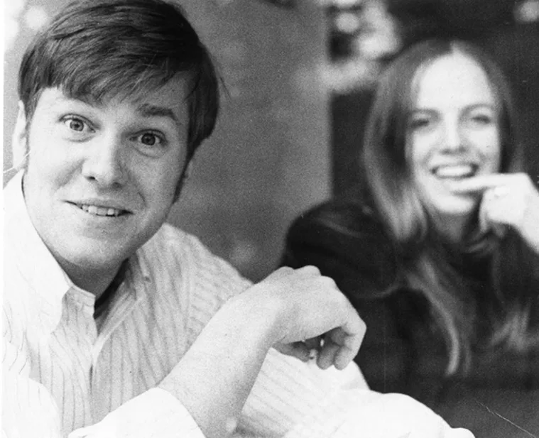 Two people are smiling, one in the foreground looking at the camera, and one in the background slightly blurred.