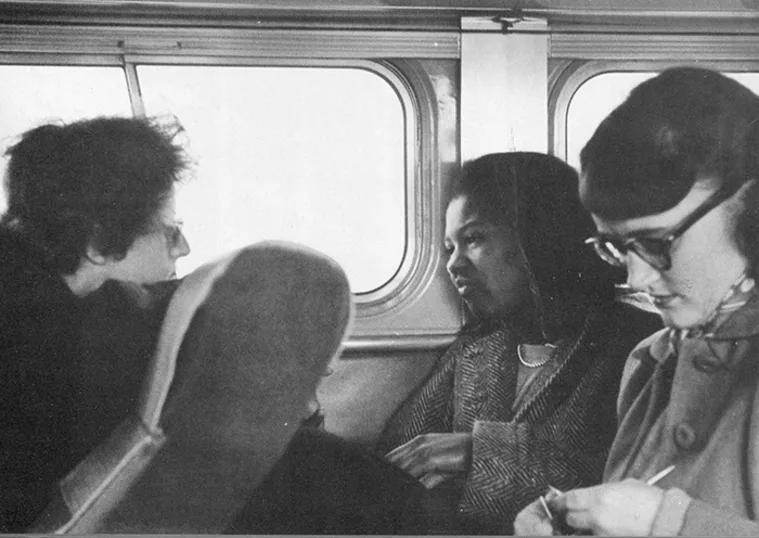 Three people seated on a bus, engaged in conversation. Two are facing each other, while the third writes notes.