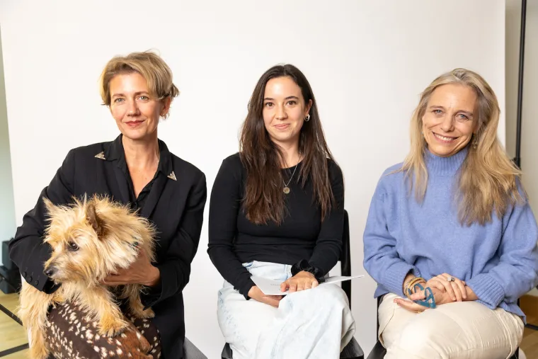 Three people sitting in a row indoors against a plain background. One person holds a small fluffy dog.
