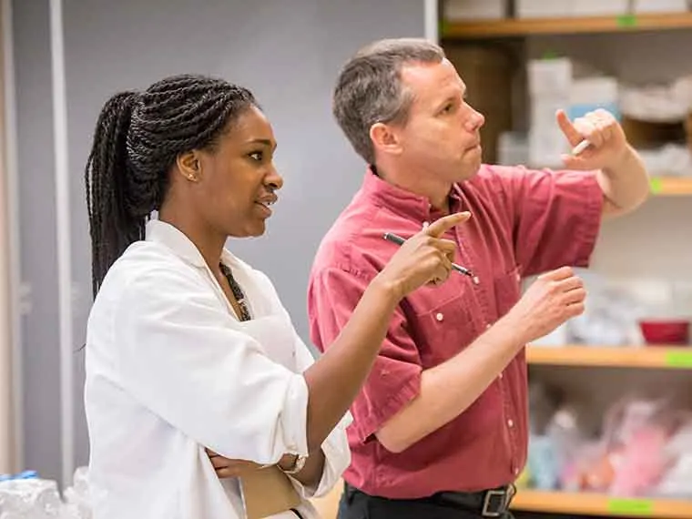 Two people in a discussion, wearing a white coat and a red shirt, gesturing with hands, standing in a room with shelves in the background.