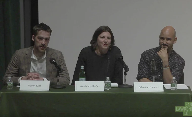 Three individuals seated at a panel discussion table with microphones and name tags in front of a wall, engaged in conversation.