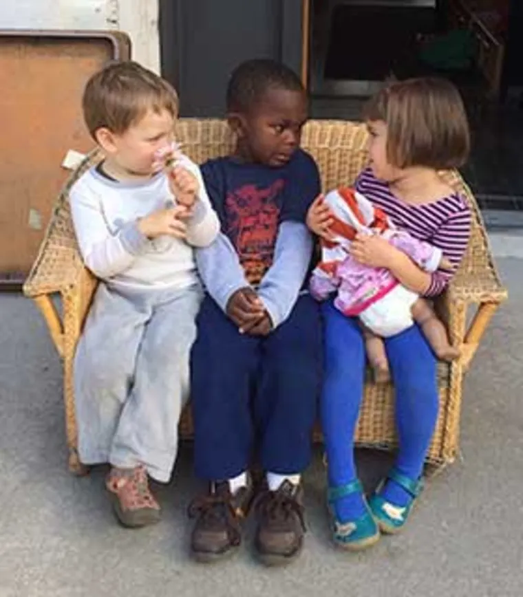 Three children sitting on a wicker bench. The one on the left is eating, the middle one looks at the right, and the one on the right holds a doll.