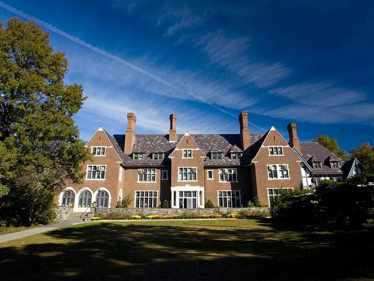Large brick mansion with multiple chimneys under a clear blue sky, surrounded by trees and grass.