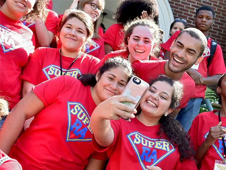 Group of smiling people in red "Super RA" shirts taking a selfie, standing closely together outside a building.