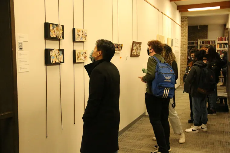 People wearing masks observe artwork in a gallery with pieces displayed on a white wall. Some hold backpacks while viewing.