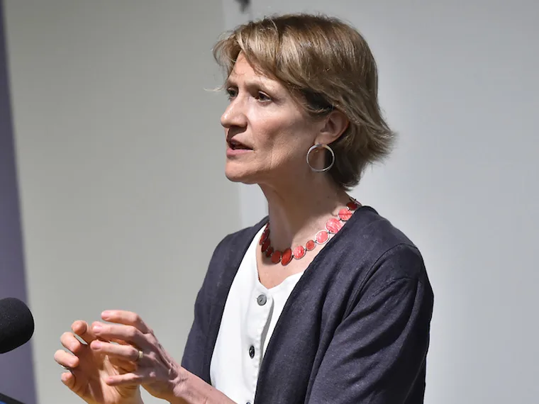 Person with short hair speaking, wearing a red necklace and hoop earrings, gesturing with one hand.