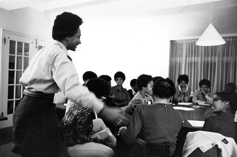 Person standing and smiling in front of seated group around a table in a room with bright lighting, engaging in a lively discussion.