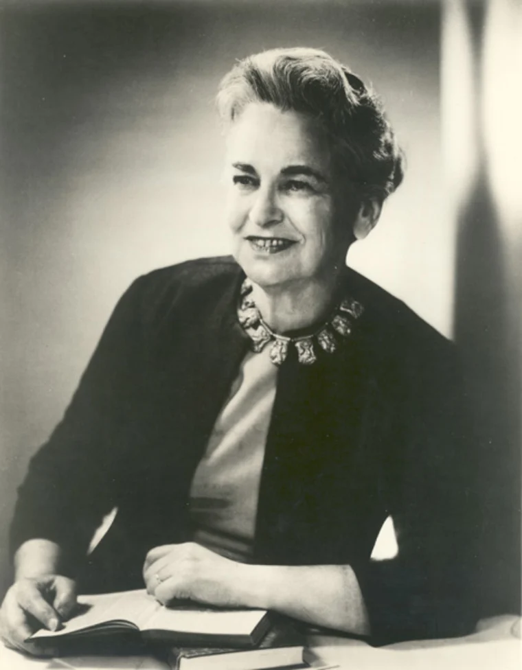 Person sitting at a table, smiling, with a book open in front. Wearing a dark jacket and a patterned necklace. Black and white photograph.