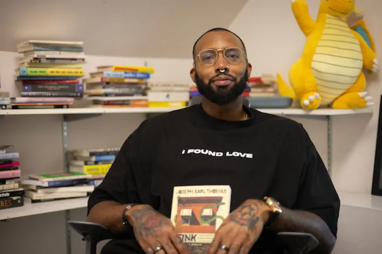 Person sitting in a chair holding a book, surrounded by bookshelves and a plush dragon toy on the shelf behind.