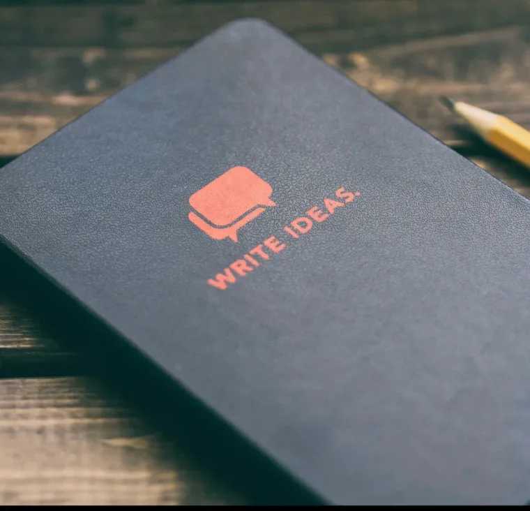 A notebook cover with the words "Write Ideas" and a speech bubble logo. A pencil lies beside it on a wooden surface.