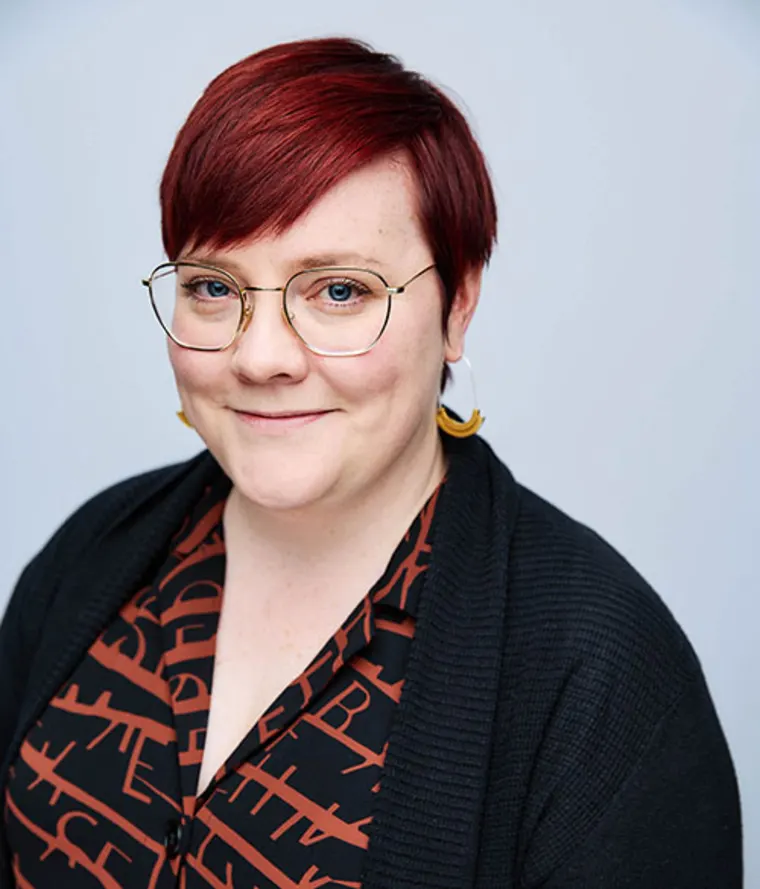 Person with short red hair and glasses, wearing a patterned shirt and black cardigan, smiles at the camera against a light background.