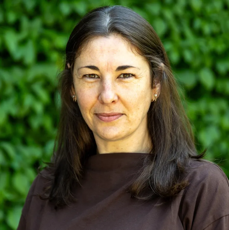 Person with long dark hair and a brown top stands in front of a leafy green background, looking at the camera.