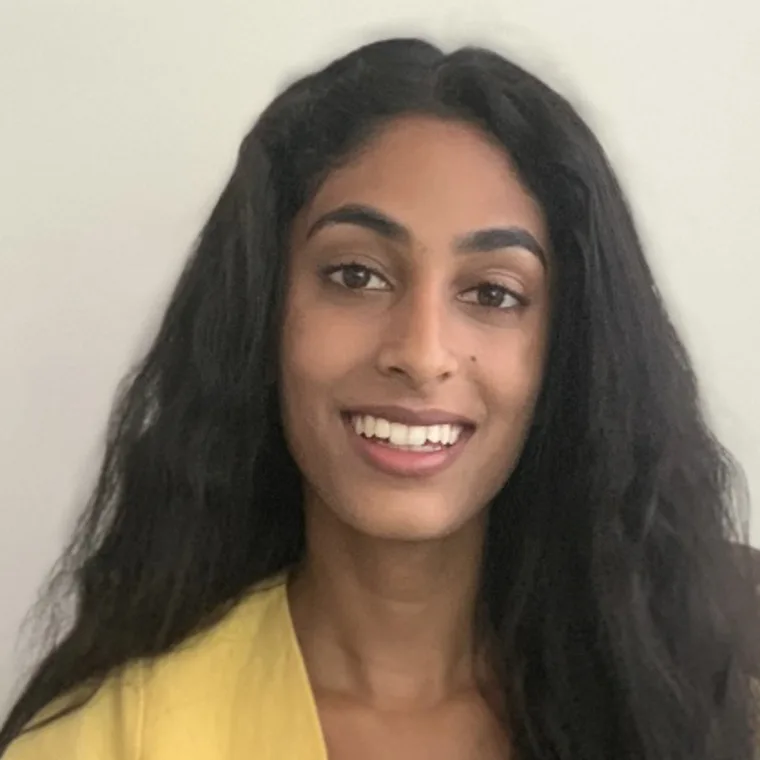 A person with long dark hair smiles warmly, wearing a yellow top, against a plain background.