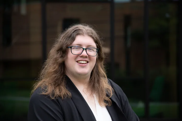 Person with long hair and glasses smiling in front of a blurred building background.