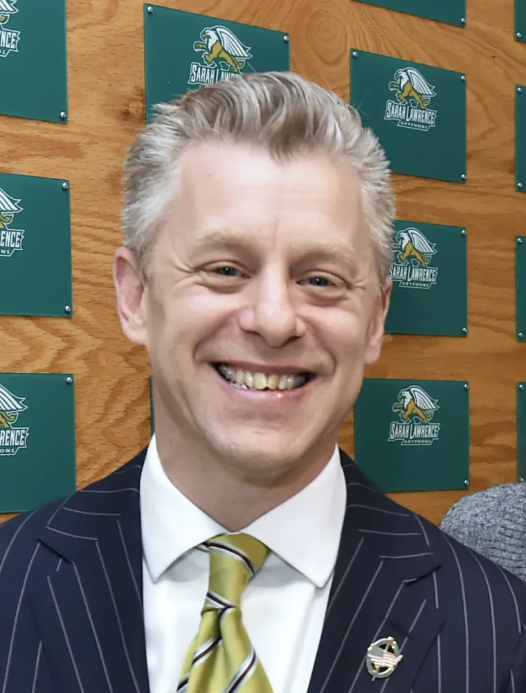 Smiling individual in a pinstripe suit and yellow-green tie, standing in front of a wall with Sarah Lawrence College logos.