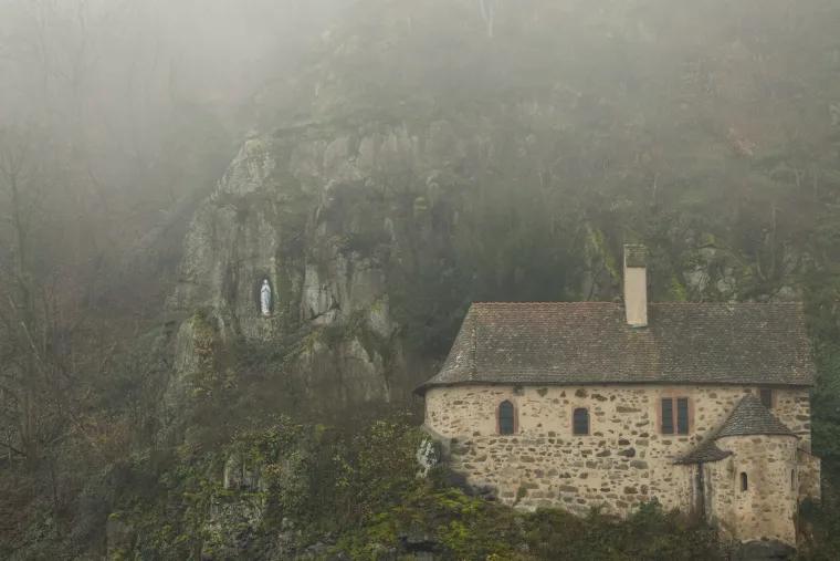Stone house beside rocky cliff with a small statue in a niche, surrounded by misty, forested landscape.