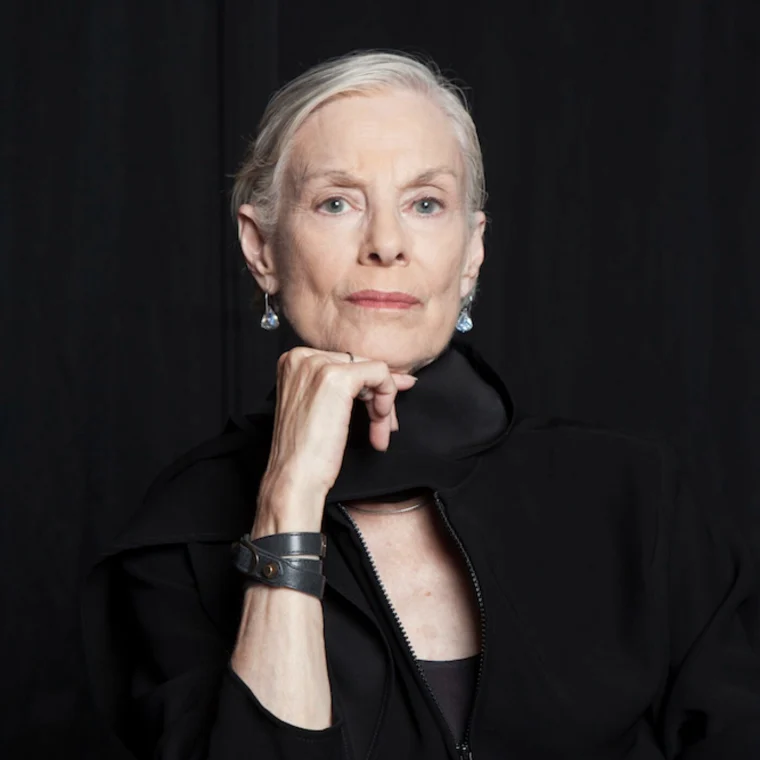 Person with short white hair, wearing earrings and a black outfit, rests chin on hand against a dark background.