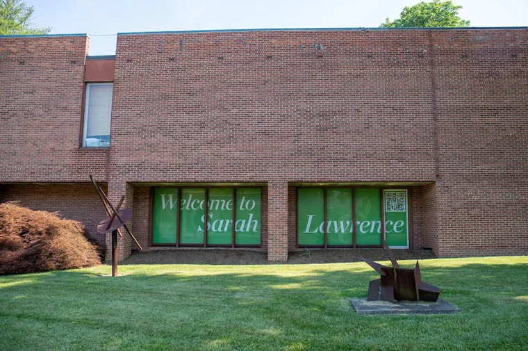 Brick building with "Welcome to Sarah Lawrence" on windows. Two modern sculptures on grassy lawn in front.