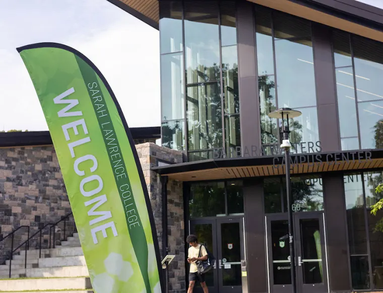 Green flag reads "Sarah Lawrence College Welcome" near a modern building labeled "Barbara Walters Campus Center." A person walks nearby.