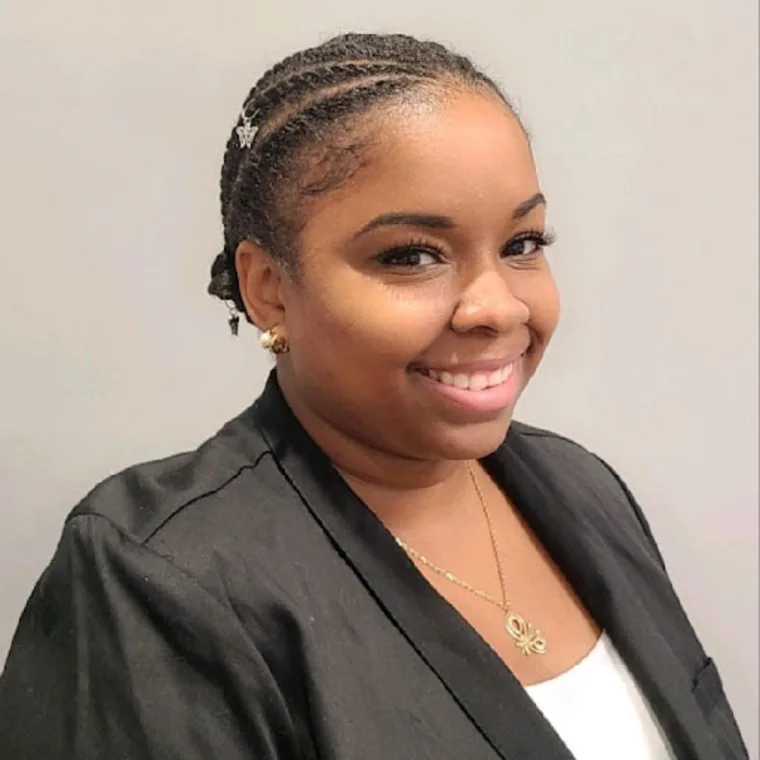 Person smiling, wearing braids with decorative clips, gold jewelry, black blazer, and white shirt against a plain background.