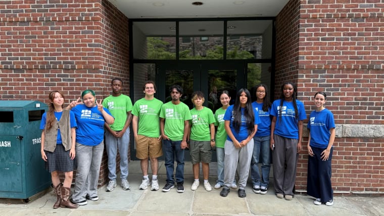 Group of ten individuals posing outside a brick building entrance, wearing blue and green shirts, some smiling, next to a trash bin.