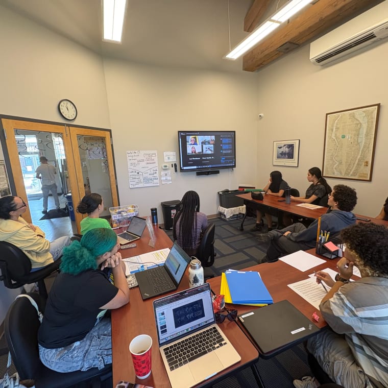 People sitting at tables in a conference room, watching a presentation on a wall-mounted screen. Laptops and papers are on the tables.