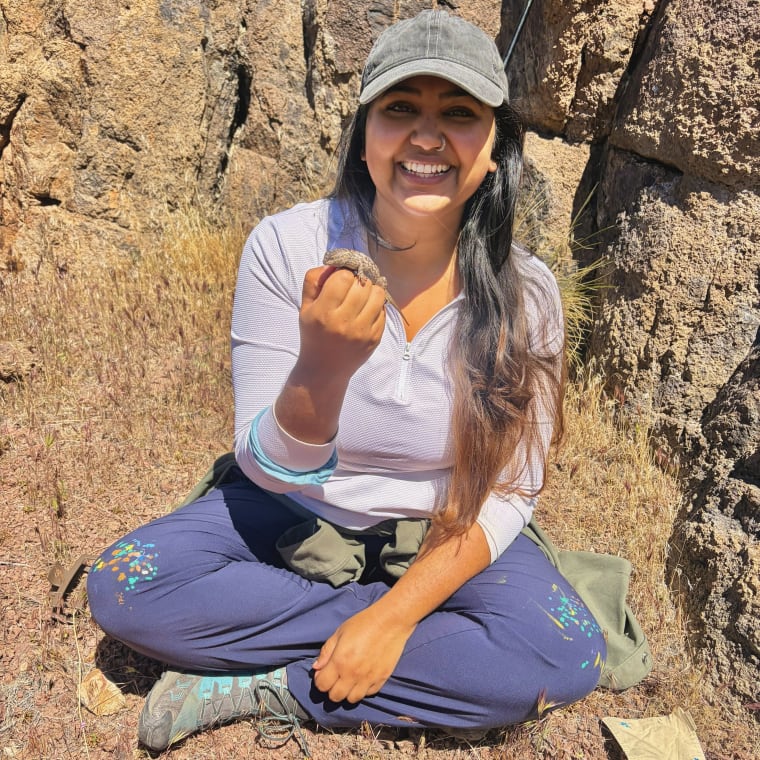 Person sitting on the ground near rocks, smiling, holding a small animal. Wearing a cap, white top, and blue pants.