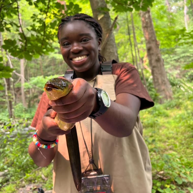 Person smiling and holding a fish with both hands in a forest setting, wearing a watch and colorful beaded bracelet.