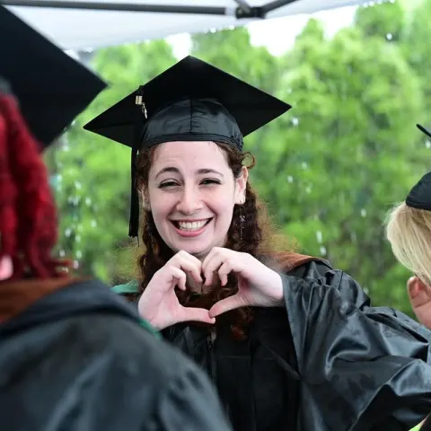 Graduate in cap and gown smiling and making a heart shape with hands, standing under a canopy with blurred green background.