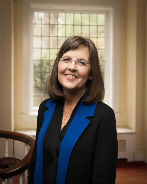 Person with shoulder-length hair smiling, wearing glasses and a black jacket with blue lapels, stands in a room with a large window and a staircase.
