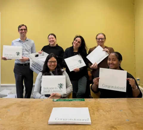 Six people smiling, holding "Welcome to Sarah Lawrence" and "Congratulations" signs in an office setting with a yellow wall.